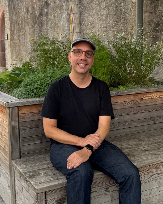 Gernot Poetsch in a cap, glasses and a black T-shirt, smiling, on a wooden bench in front of an old stone wall