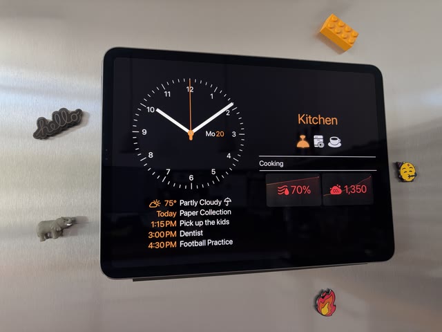 A photo: an iPad held to a stainless-steel fridge door, fridge magnets around it, showing a large clock and a kitchen’s dashboard