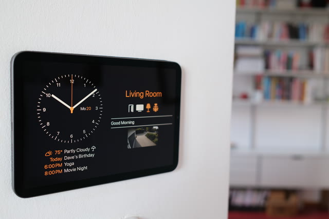 A photo: an iPad mounted flat on a white wall, showing a large clock and a living room’s dashboard, a bookshelf blurred behind it